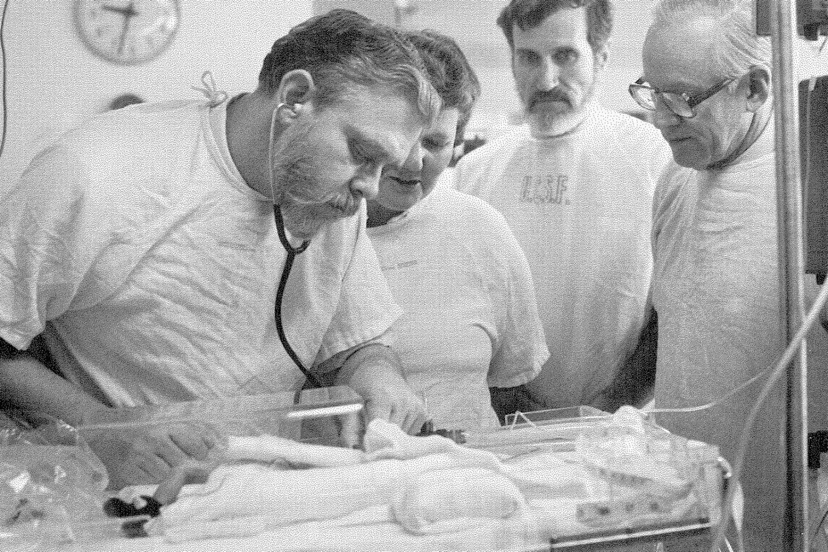 Four doctors examine a small baby in an incubator in a black-and-white photo