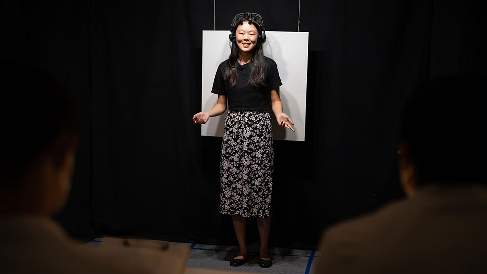 Veronica Then speaks in front of 2 people in a dark lab, standing in front of a white screen, wearing an electronic measuring device on her head.
