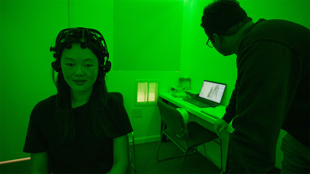 A student wearing a measuring device on her head sits in a lab bathed in green light