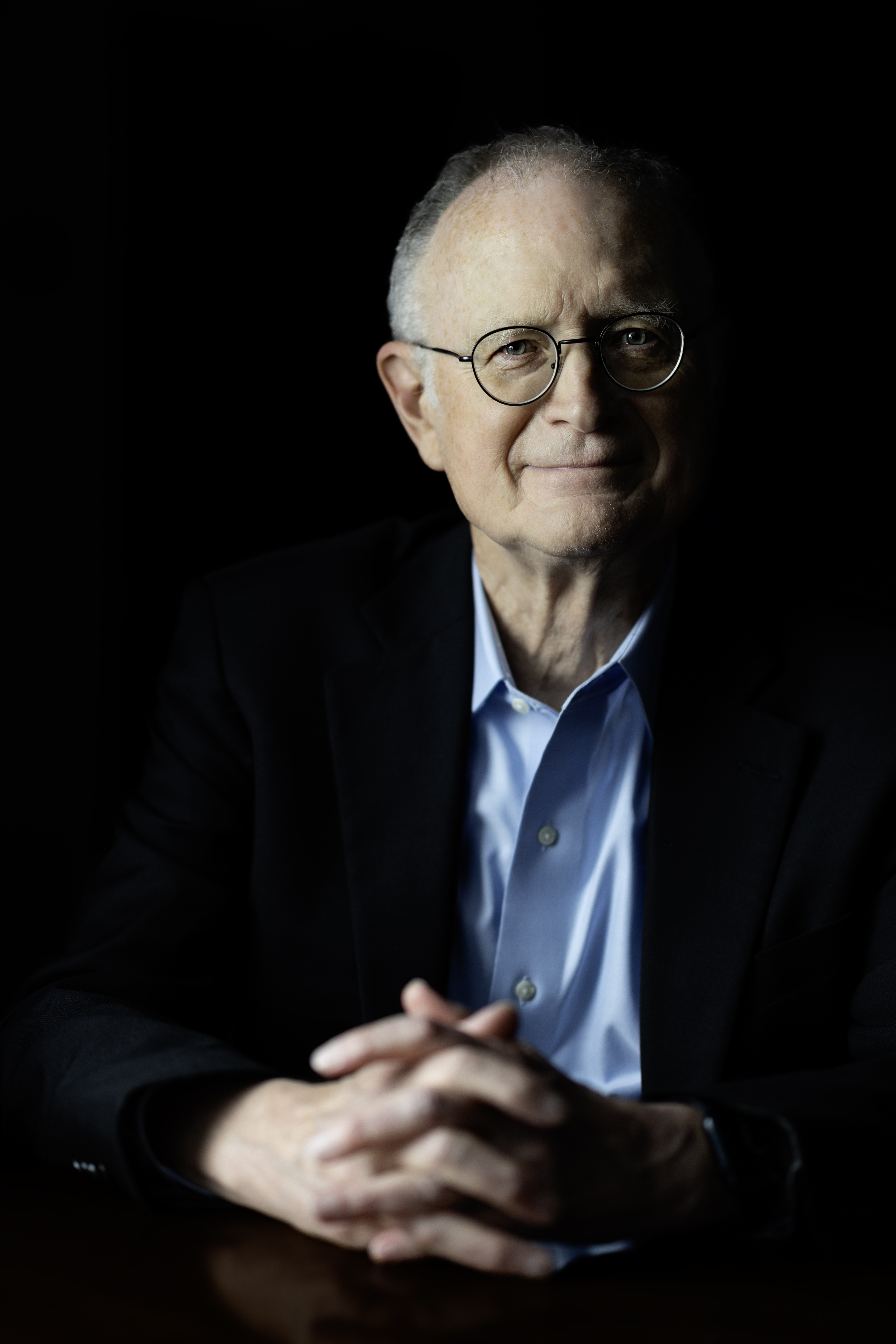 A man wearing glasses smiles at the camera, hands folded