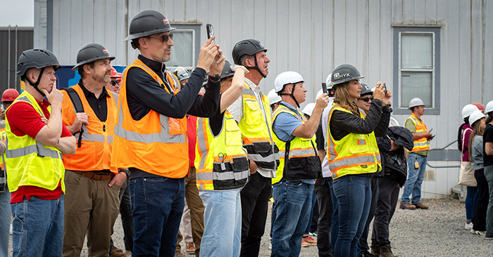 A group of people wearing safety vests and hard hats watching something in the distance, some with their phones out to take videos