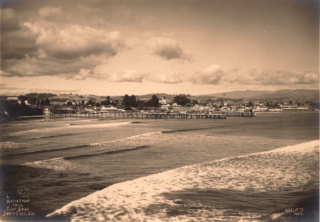 A sepia historical photograph of the Santa Cruz waterfront, with a set of waves rolling in