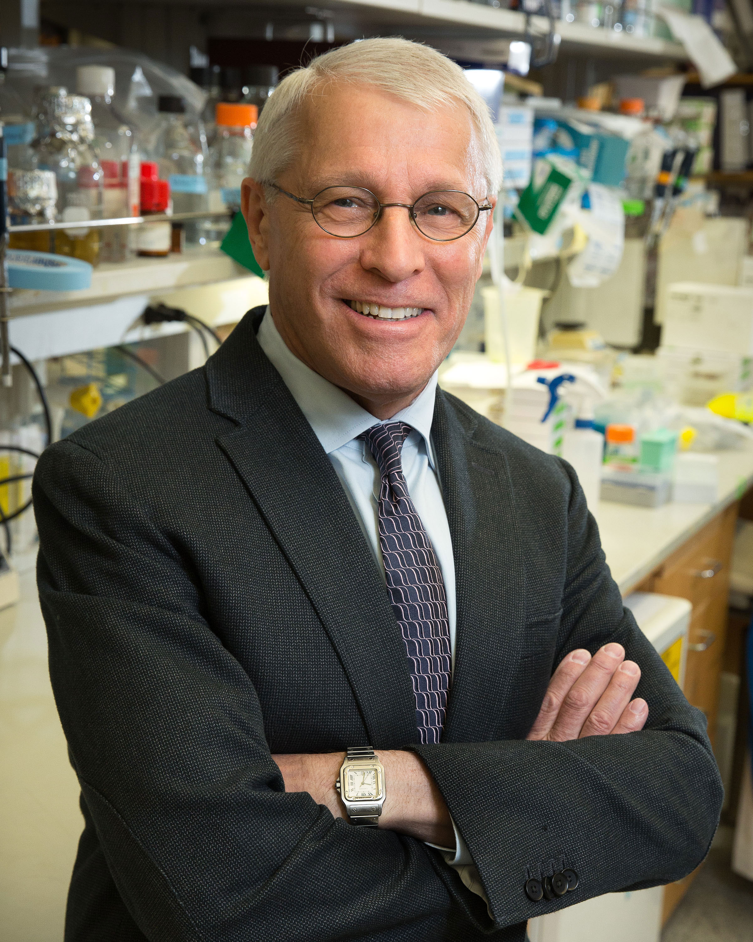 Owen Witte, wearing a suit, smiles with his arms crossed over his chest in front of a lab bench