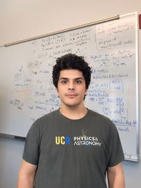 Mohamed Shalby, wearing a UCR Physical Astronomy t-shirt, looks at the camera while standing in front of a white board covered with calculations