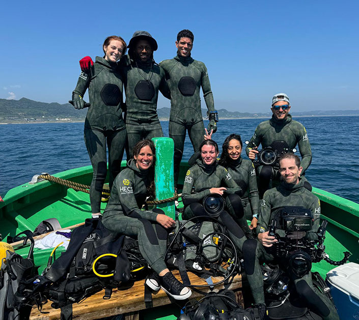 A group of men and women in scuba suits with underwater cameras on an ocean boat.