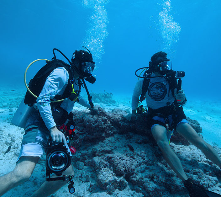 Two men in scuba gear on the ocean floor with cameras and a video filming device in hand.