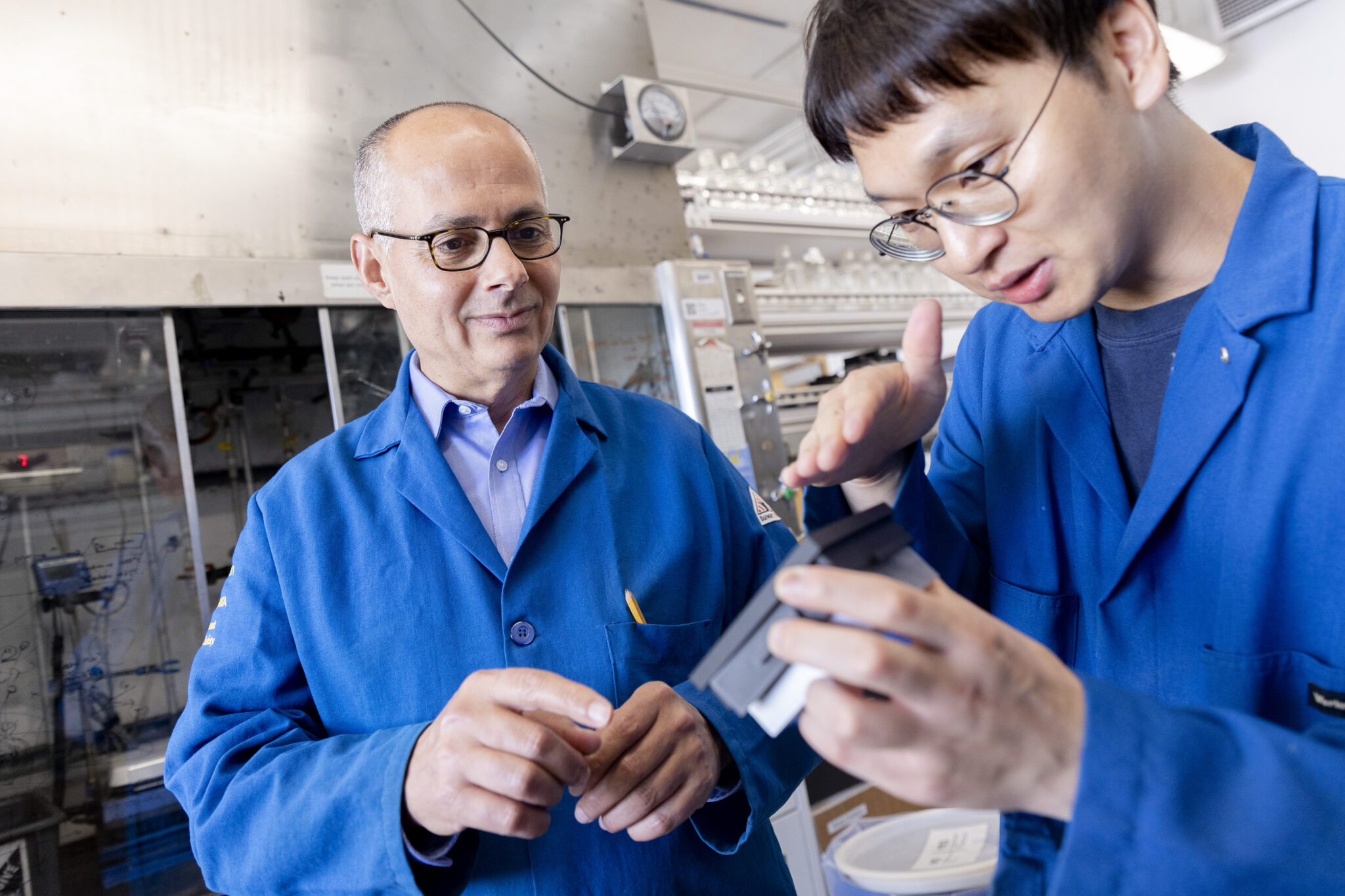 A man in glasses and a blue lab coat watches a young man hold an object and gesture at it