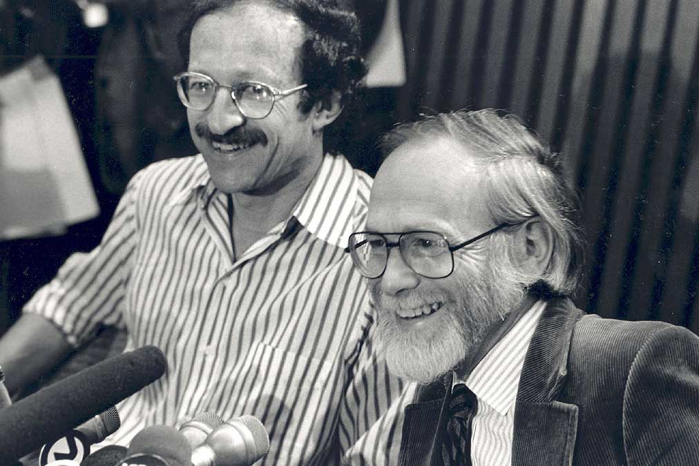 Two men in a black and white photo smile while seated together in front of a mic