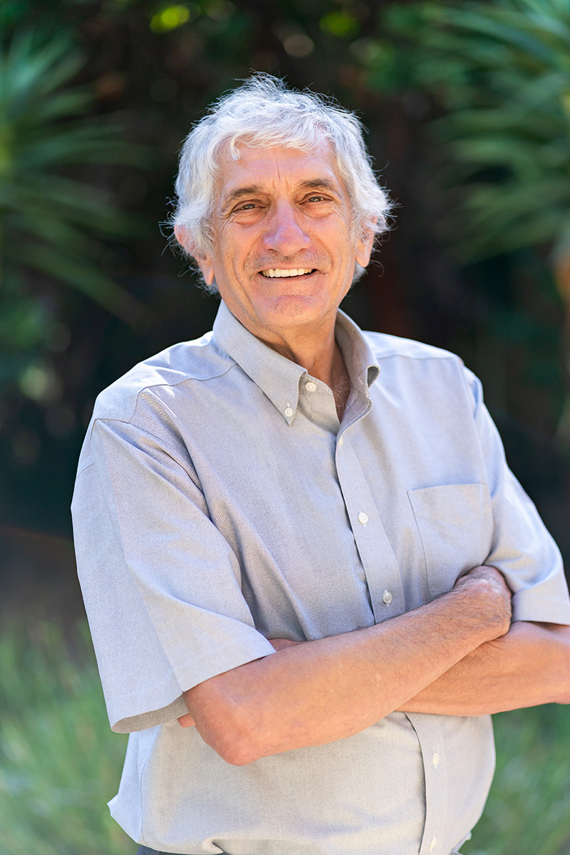 John Martinis smiles at the camera with his arms crossed over his chest, standing in front of vegetation.