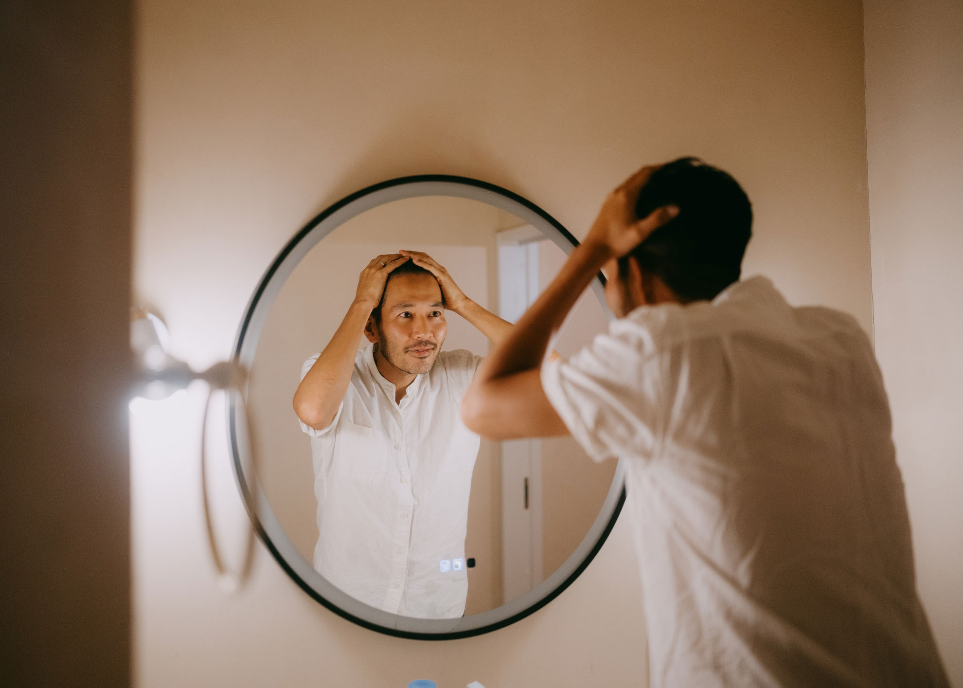 A man with dark, short hair looks into a circular mirror, with his hands on his head