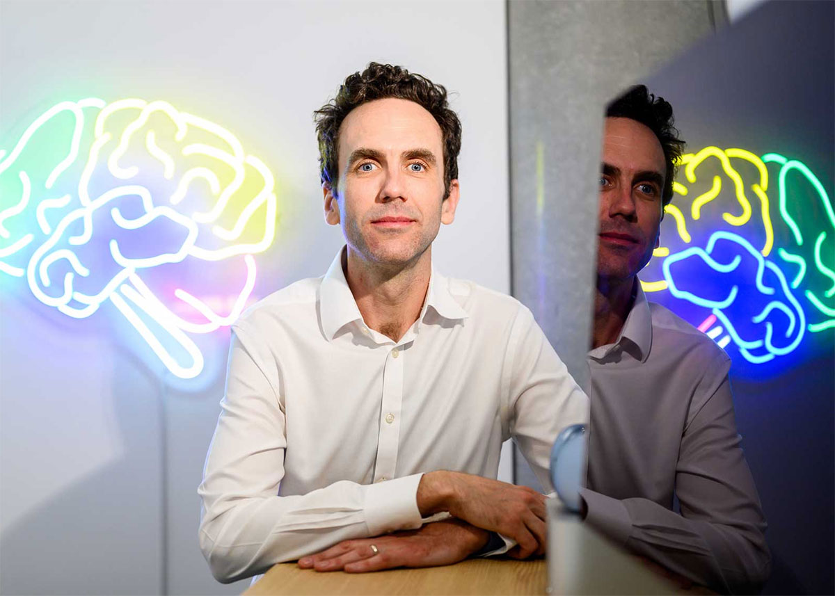 Simon Little looks at the camera, sitting in front of a large wall art that's a neon tube rendering of the human brain