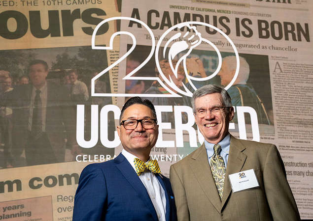 Two men in suits stand smiling in front of a large backdrop featuring the words “20 UC Merced” and old newspaper headlines reading “A Campus Is Born.” The display celebrates the 20th anniversary of the University of California, Merced.