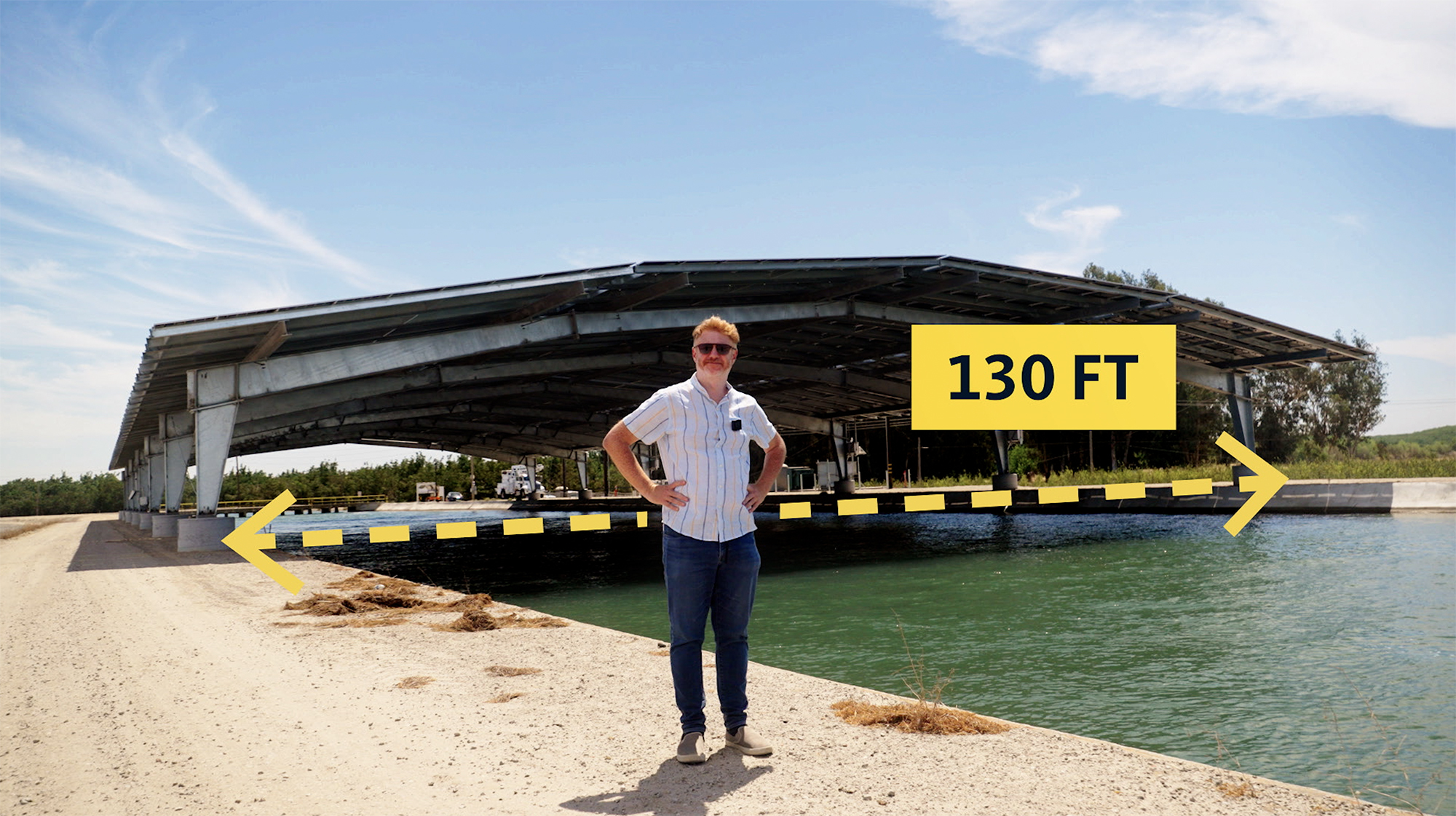 A man stands in front of a wide canopy of solar panels over a canal that has a yellow dotted line with an arrow at each end that says 130 feet
