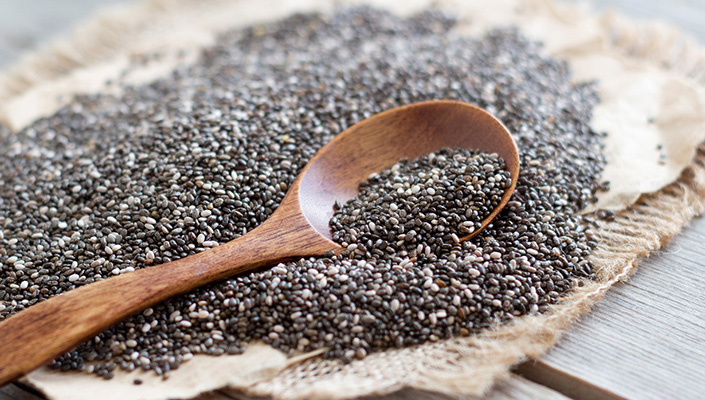 A pile of black chia seeds on a burlap patch with a wooden spoon resting on the pile
