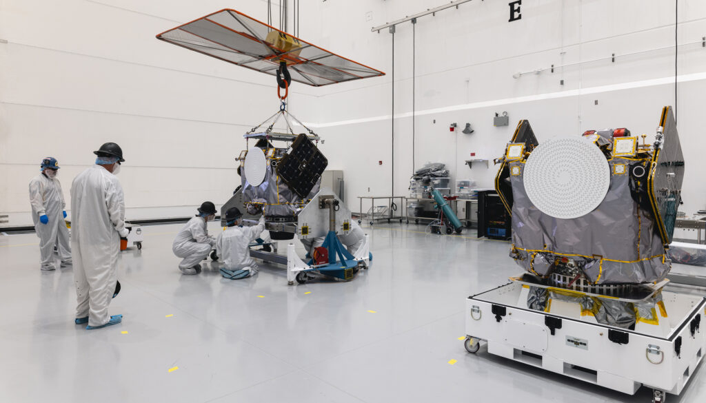 People in cleanroom suits work on spacecraft components inside a sterile assembly facility. One satellite is suspended by a crane while technicians inspect and adjust it, and another completed satellite sits nearby on a wheeled platform. The bright, spotless environment is equipped with lab tools and equipment along the walls.