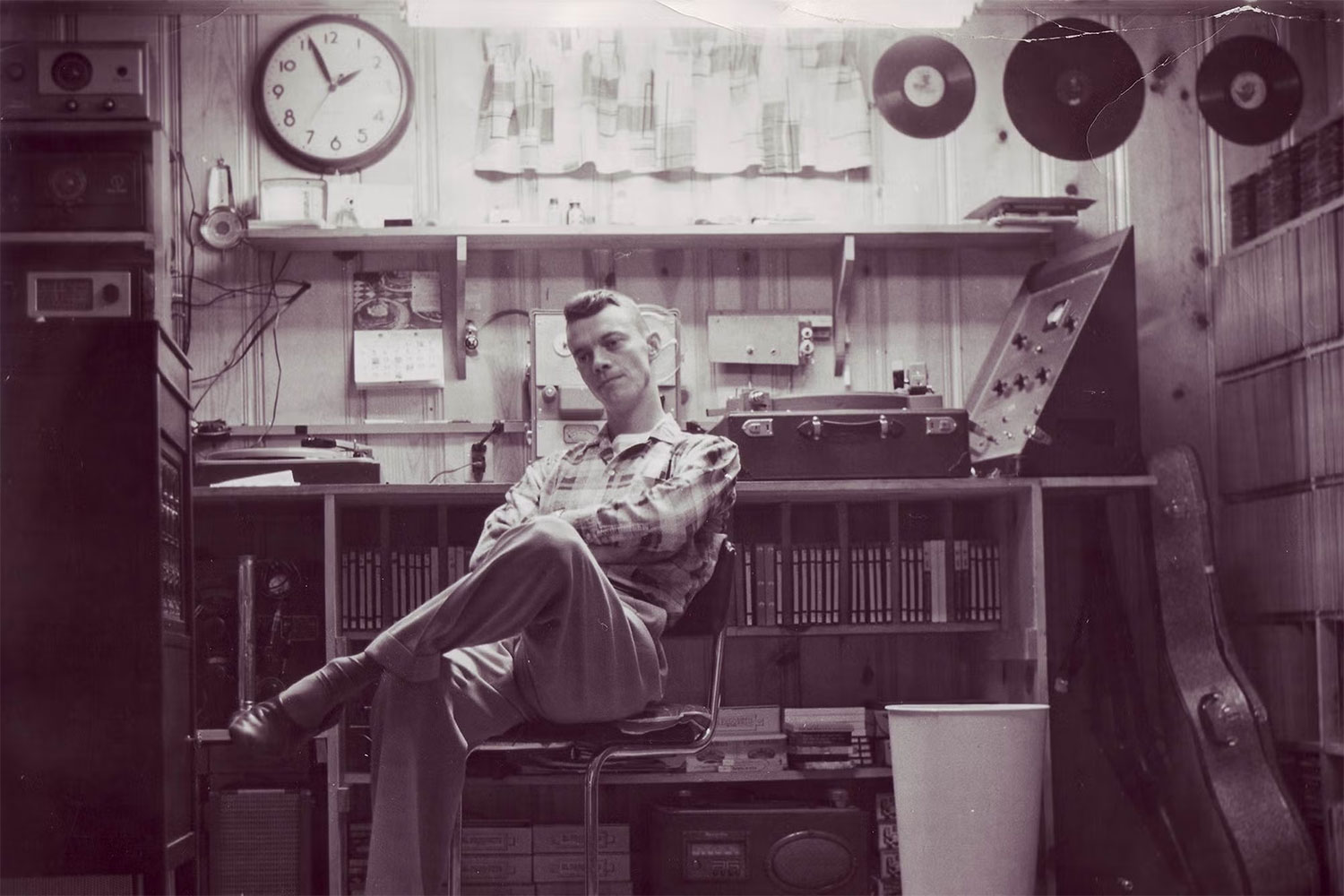 Sepia toned image of a man with a 1950s haircut and attire in a music studio, in a chair with one leg crossed over the other knee.