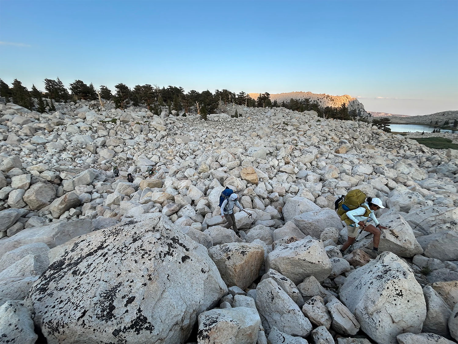 Two hikers in backpacks with poles navigate a granite boulder field.
