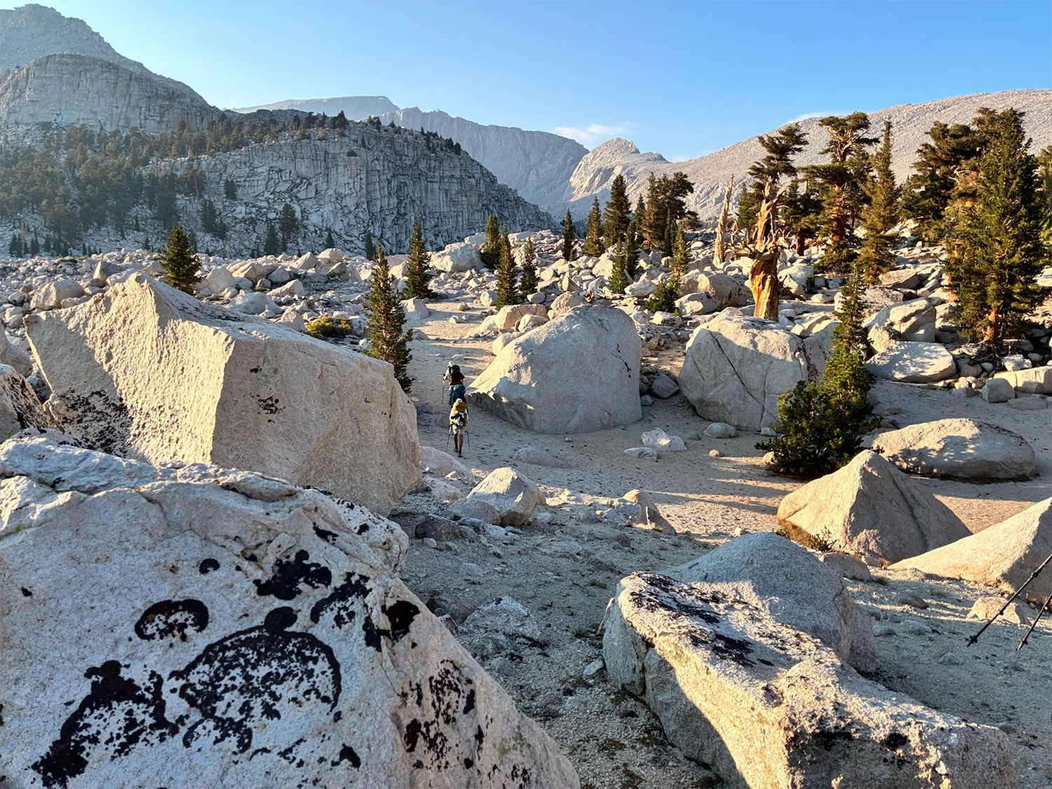 Two people walk through a granite boulder landscape with scrubby coniferous trees.