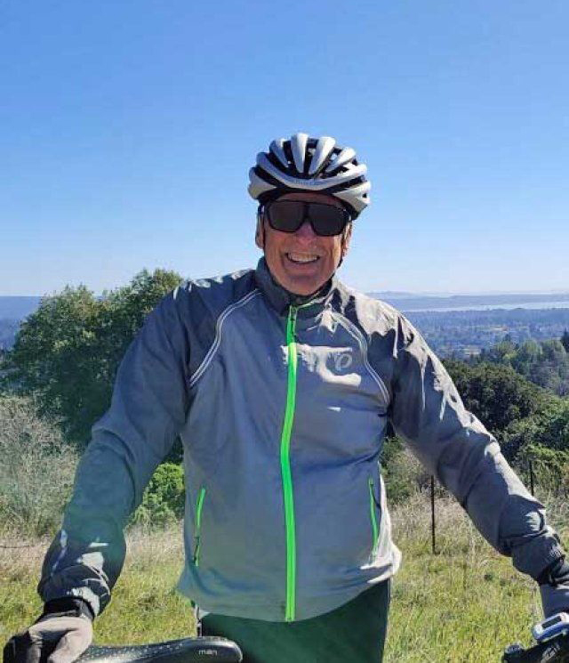 A man wearing a bike helmet and a jacket stands holding a mountain bike on a bright day