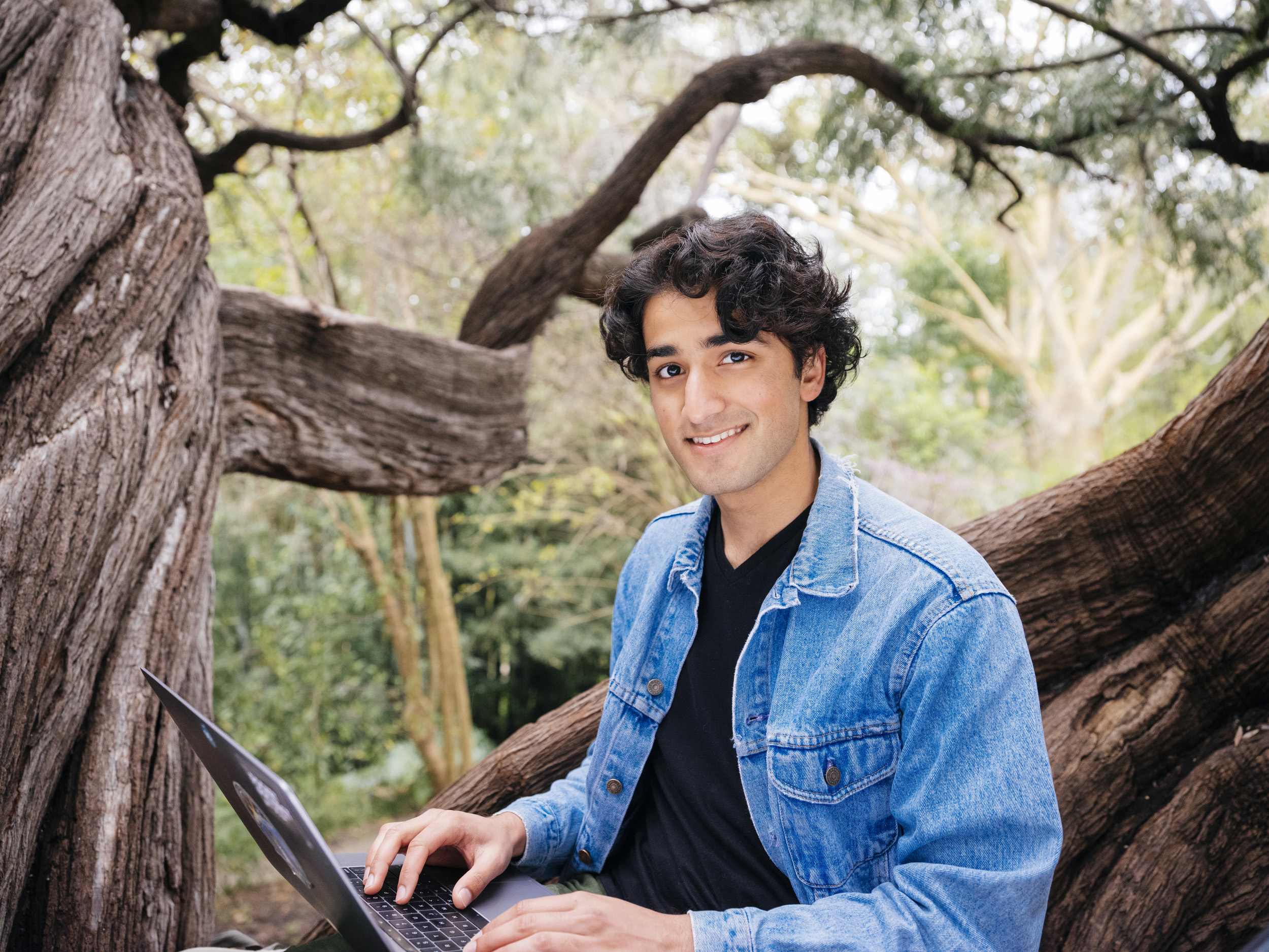 A young man in a jean jacket uses a laptop while sitting in a tree and smiling at the camera