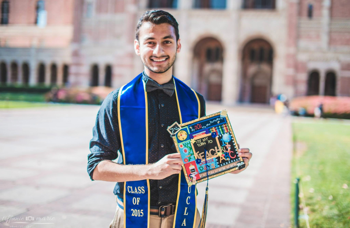 A man with a UCLA Class of 2015 graduation stole standing in front of a UCLA building, smiling and holding a mortarboard decorated with the words "It's my turn to teach"