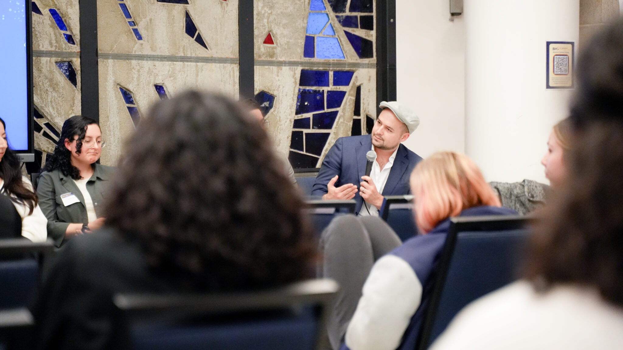 A man in a suitcoat and newsboy hat holds a microphone and gestures at an event.