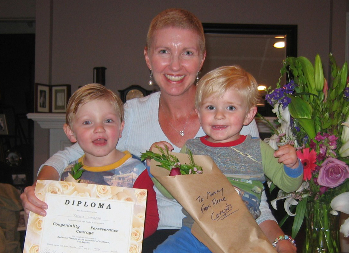 A smiling woman with very short hair holds two small, happy children, a diploma and a bouquet of flowers
