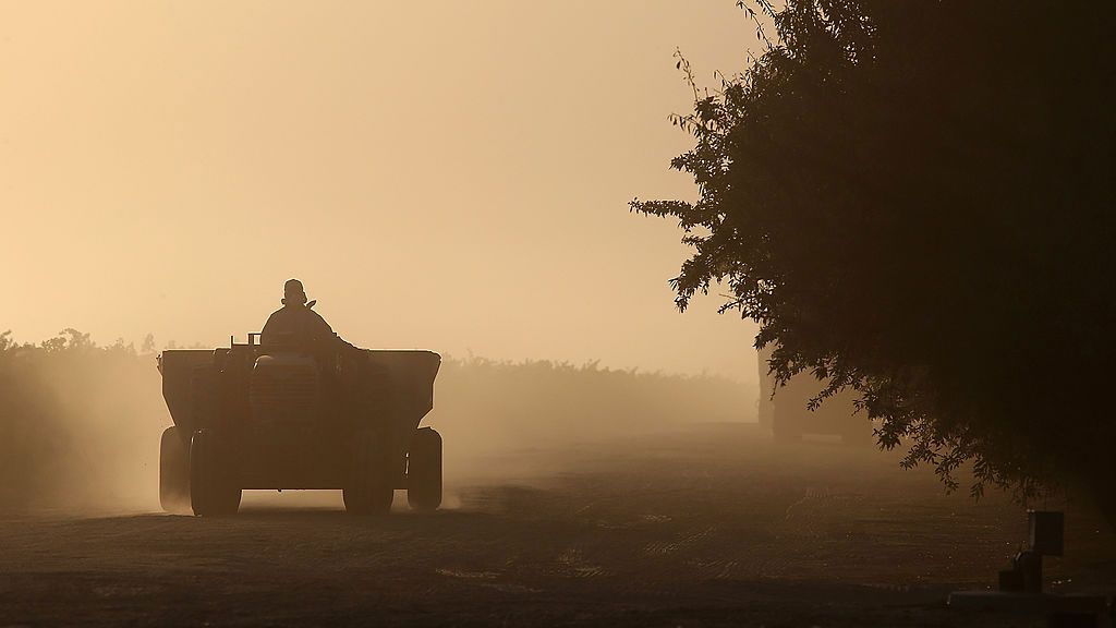 A tractor drives down a dirt road at sunset, kicking up dust that fills the air. The scene is silhouetted, with trees lining the right side of the frame and the sky glowing warm in the background.