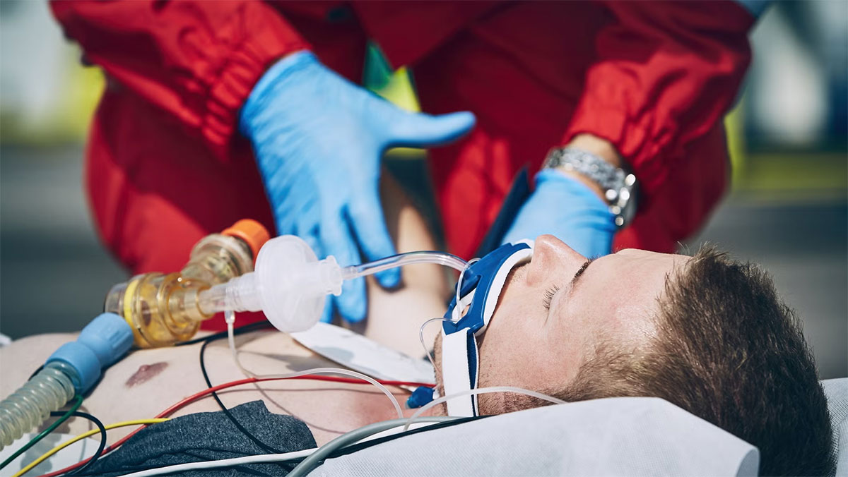 A paramedic wearing blue gloves and red protective gear provides emergency care to an unconscious man lying on a stretcher, who has a breathing tube and medical monitoring wires attached to his chest.