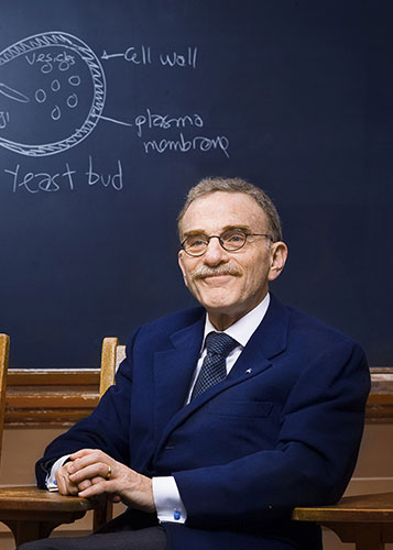 Randy Schekman wearing a blue suit and spectacles sits in a lecture classroom desk, in front of a chalkboard with a diagram of a yeast cell drawn on it, wearing a blue suit and smiling affably