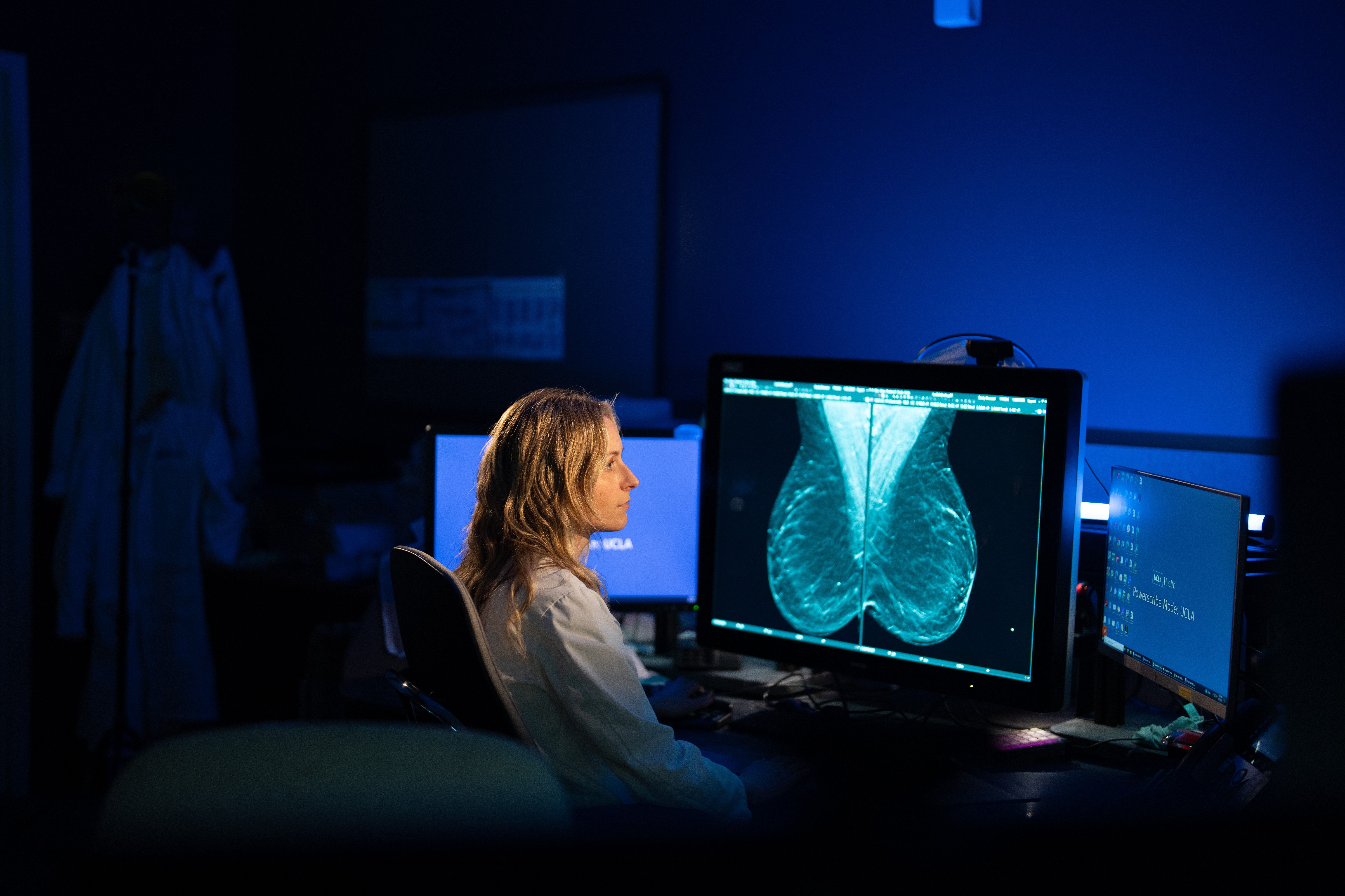 A clinician seated at a workstation in a dimly lit radiology room, viewing a detailed mammogram image on a large computer monitor, with additional screens and lab coats visible in the background.