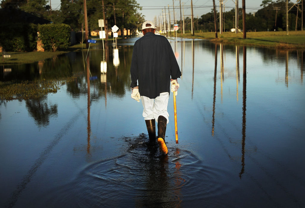 A person wearing rubber boots walks through floodwater covering a residential street, using a walking stick as utility poles and road signs reflect in the water.