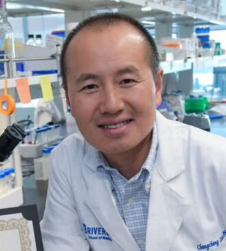 Changcheng Zhou looks up from a microscope eyepiece to smile at the camera in a lab