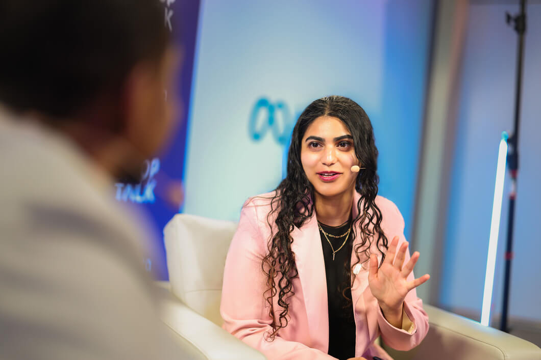 A woman with a small mic speaks to someone whose shoulder we see