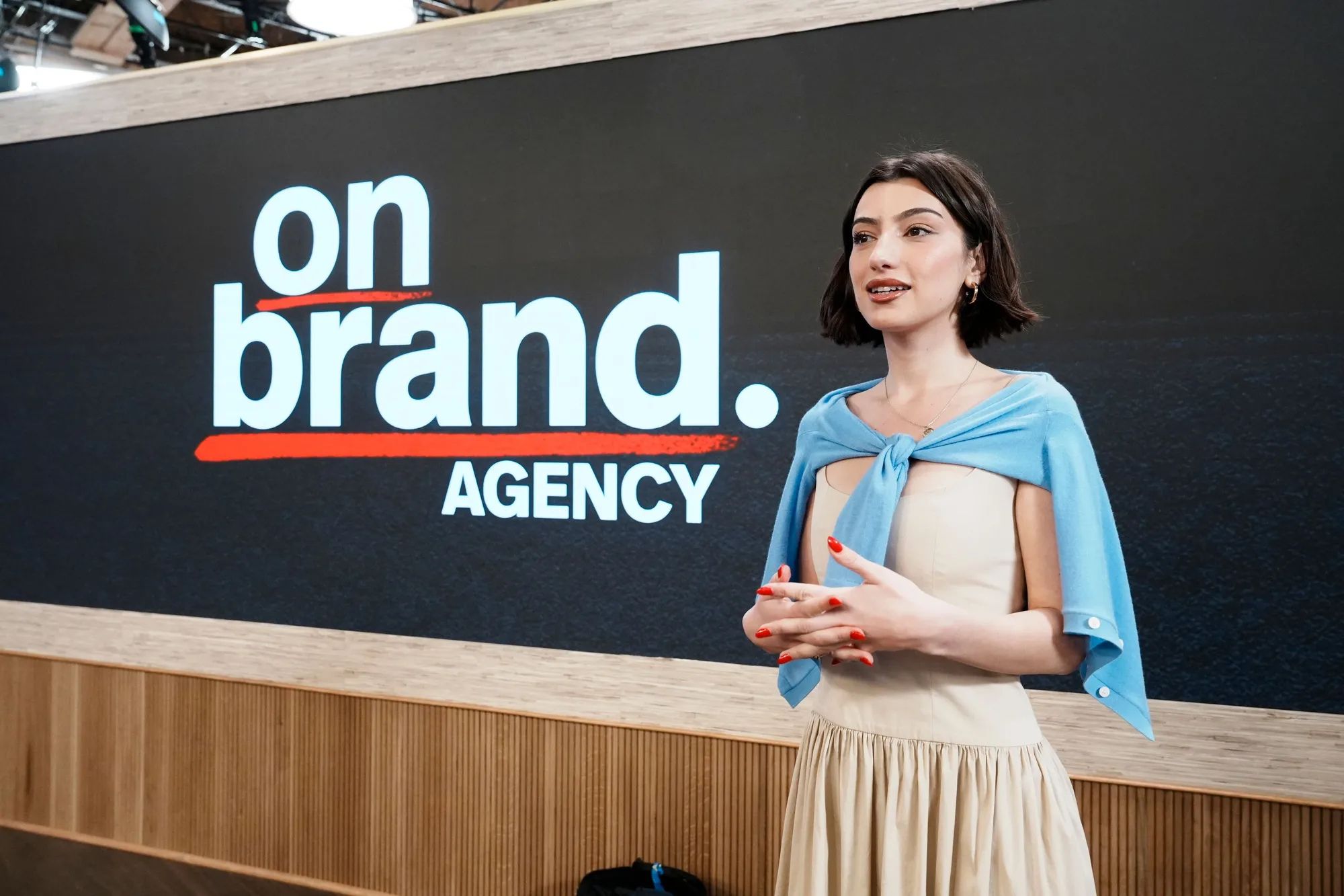A young woman with a cardigan around her shoulders stands in front of a sign that says on brand. agency