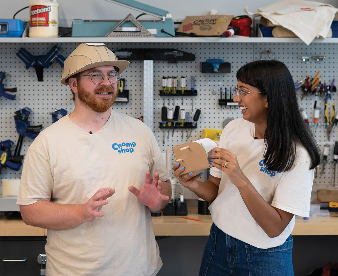 In a neat toolshed, a man in a cardboard hard hat next to a woman holding a cardboard item