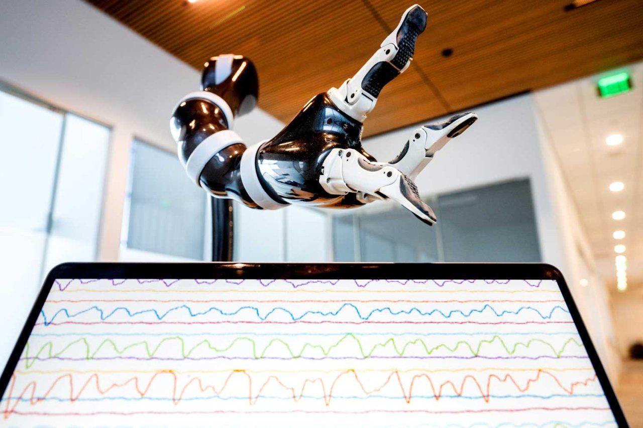 A robotic arm hovers over a computer screen showing stacks of brainwave recordings in a lab.