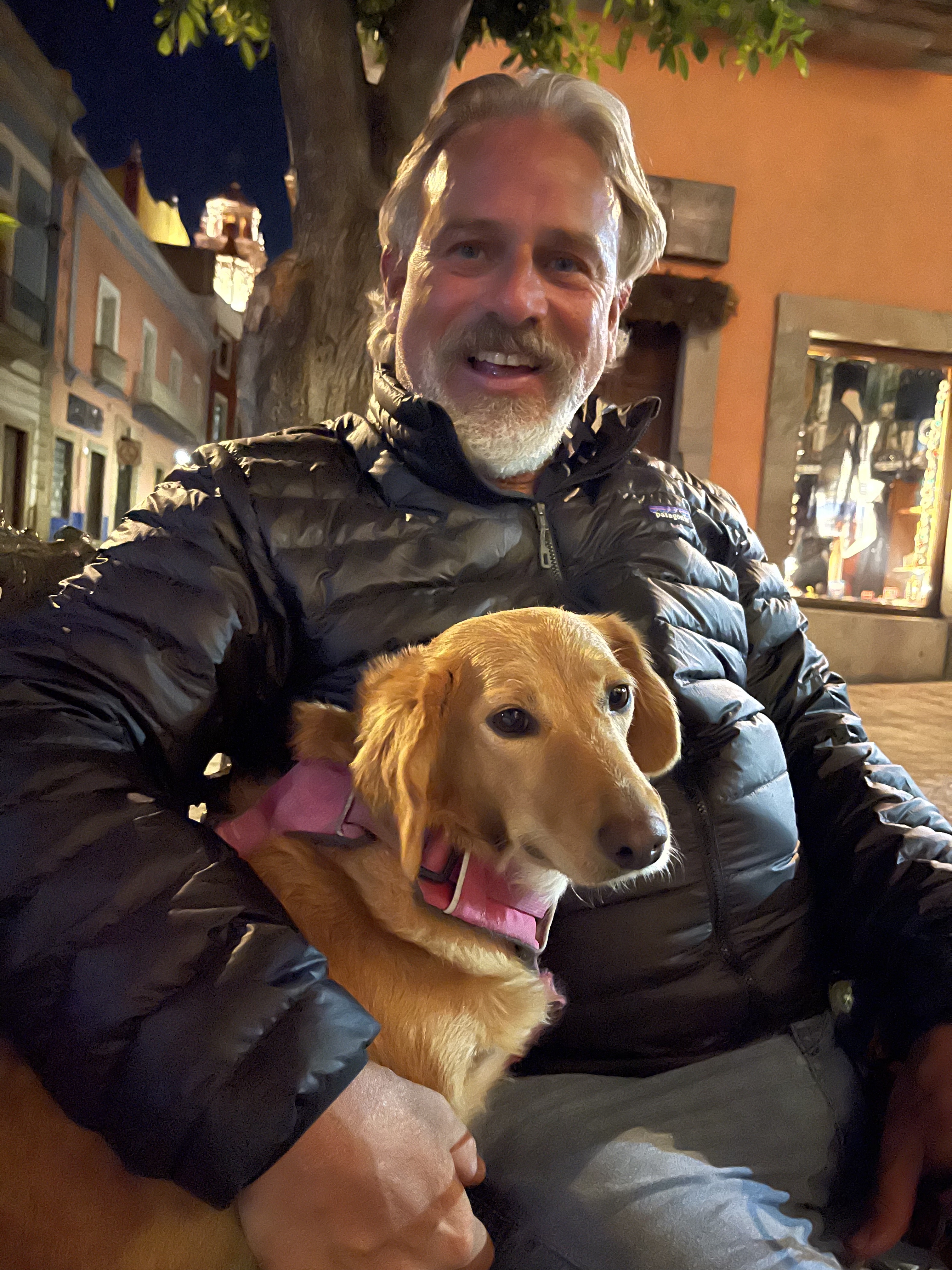 A man smiles while sitting with his dog, both looking at camera, in an Italian town at night