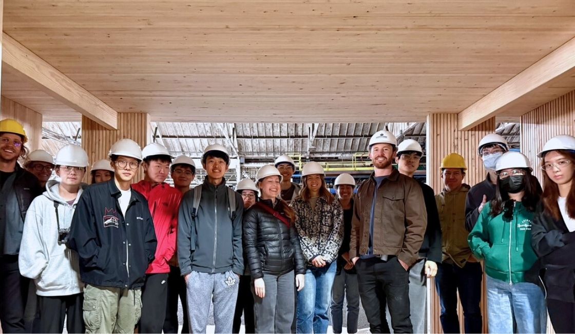 Group of people wearing hard hats standing beneath a wood ceiling and between timber columns, posing together inside a mass timber building under construction.