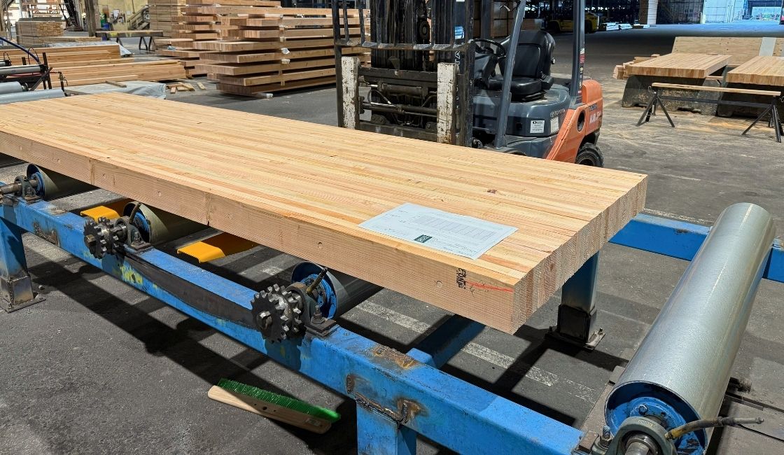 Large laminated timber panel resting on rollers inside a manufacturing facility, with stacked wood panels and a forklift visible in the background.