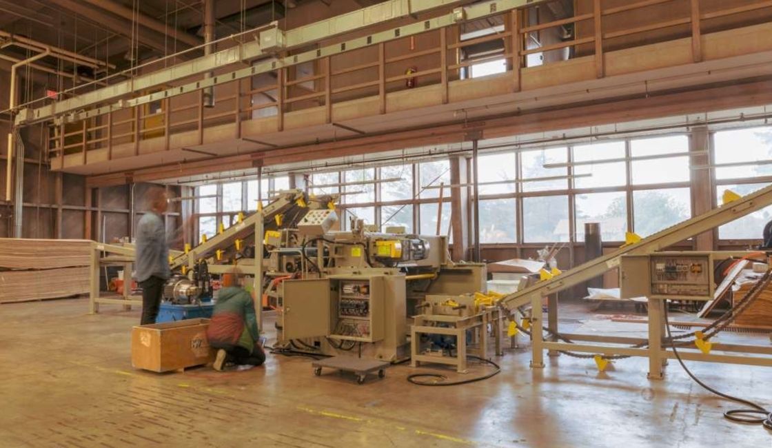 Interior of a mass timber manufacturing plant showing workers near an automated wood processing machine, with conveyor belts and stacked timber panels around the space.