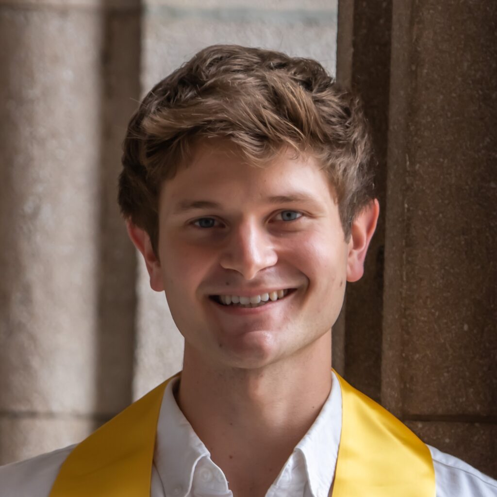 A close-cropped square portrait of Andrew Olander smiling at the camera, wearing a white button-up shirt an a yellow sash around his neck