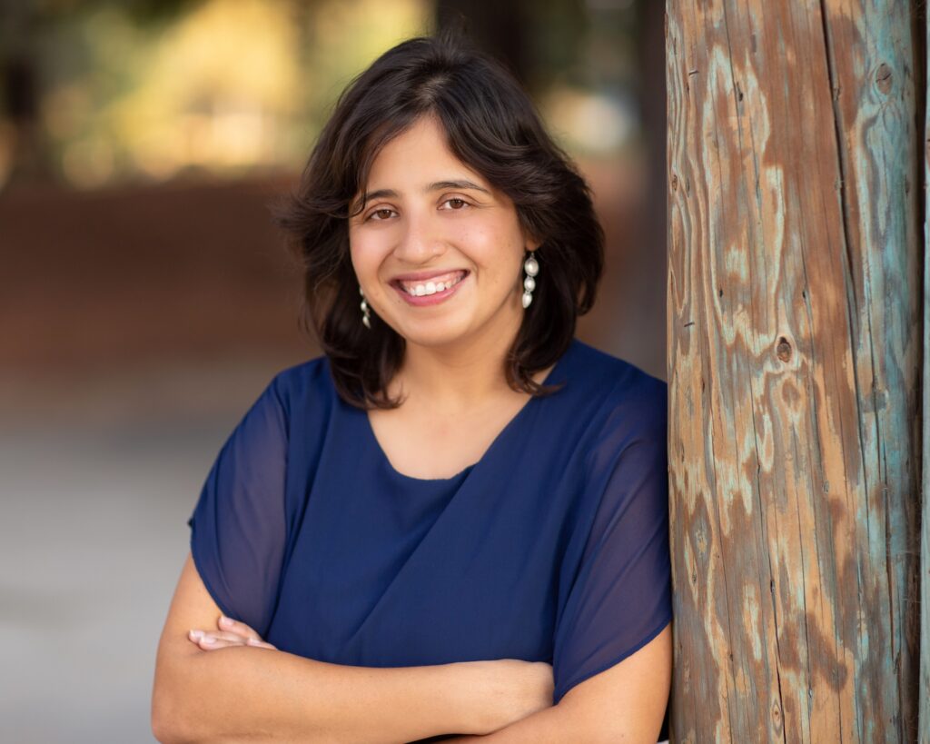 Shaheen Sikandar smiles for a portrait with arms folded over her chest, leaning against a wooden post