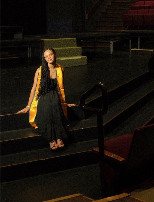 A woman in a graduation stole sits on a stage