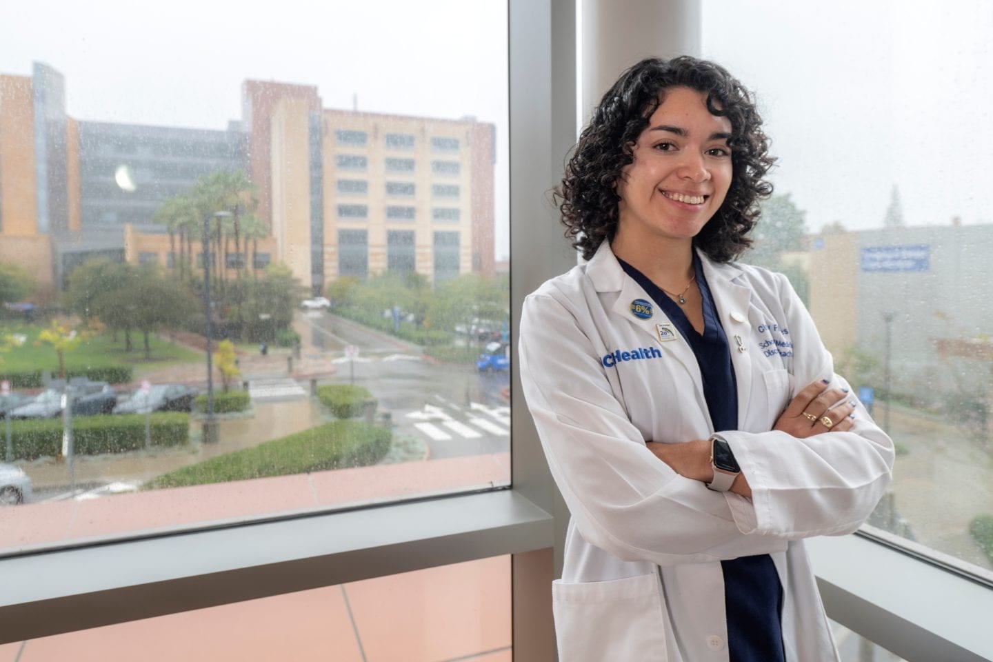 A woman with curly shoulder length hair in a white medical coat smiles with her arms crossed