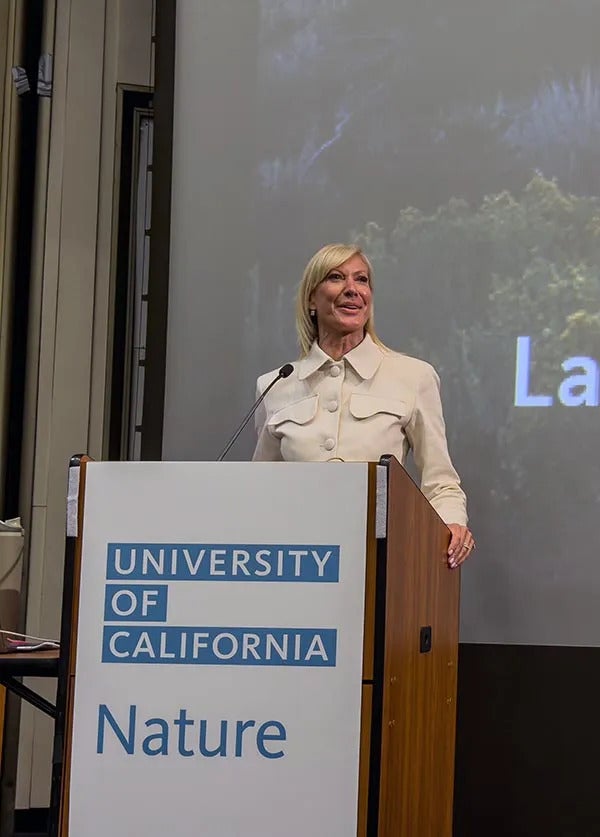 Regent Reilly in a white coat stands at a UC Nature podium, smiling
