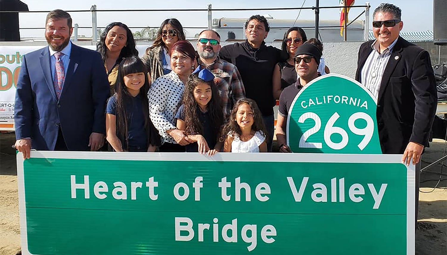 In a group photo adults and children in professional / formal wear pose behind a highway sign that says Heart of the Valley Bridge and the highway sign 269