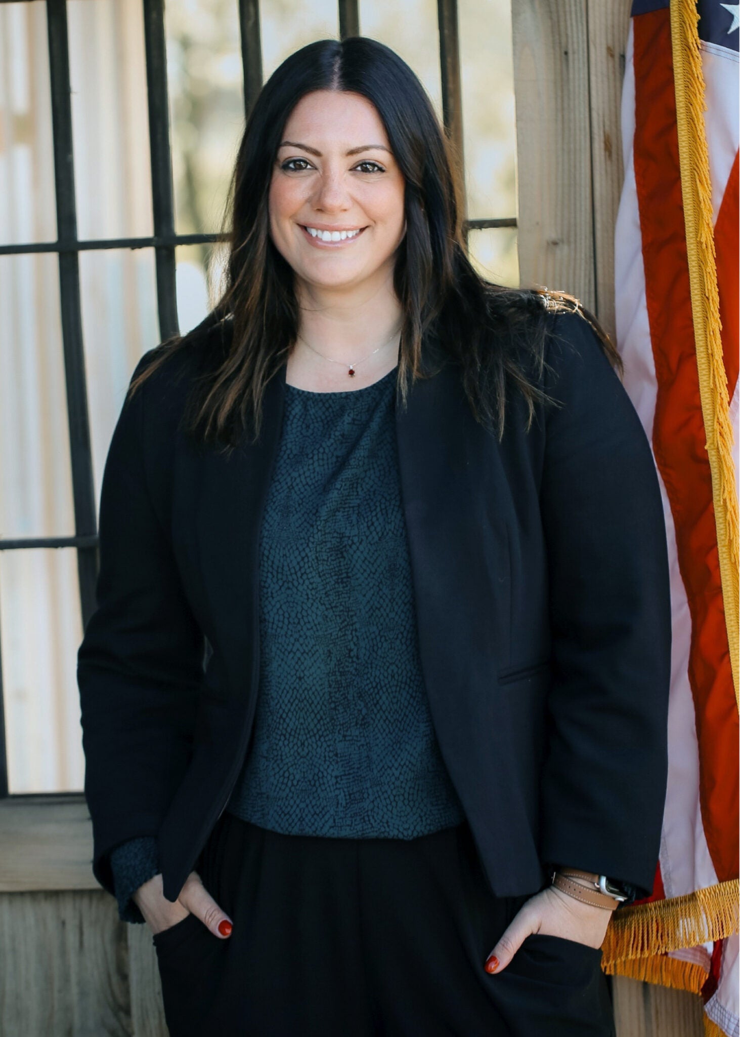 A woman with shoulder-length dark hair in a professional suit stands for a portrait next to an American flag