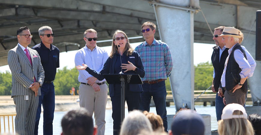 A woman speaking at a microphone in front of a solar panel canopy while a group of men listen