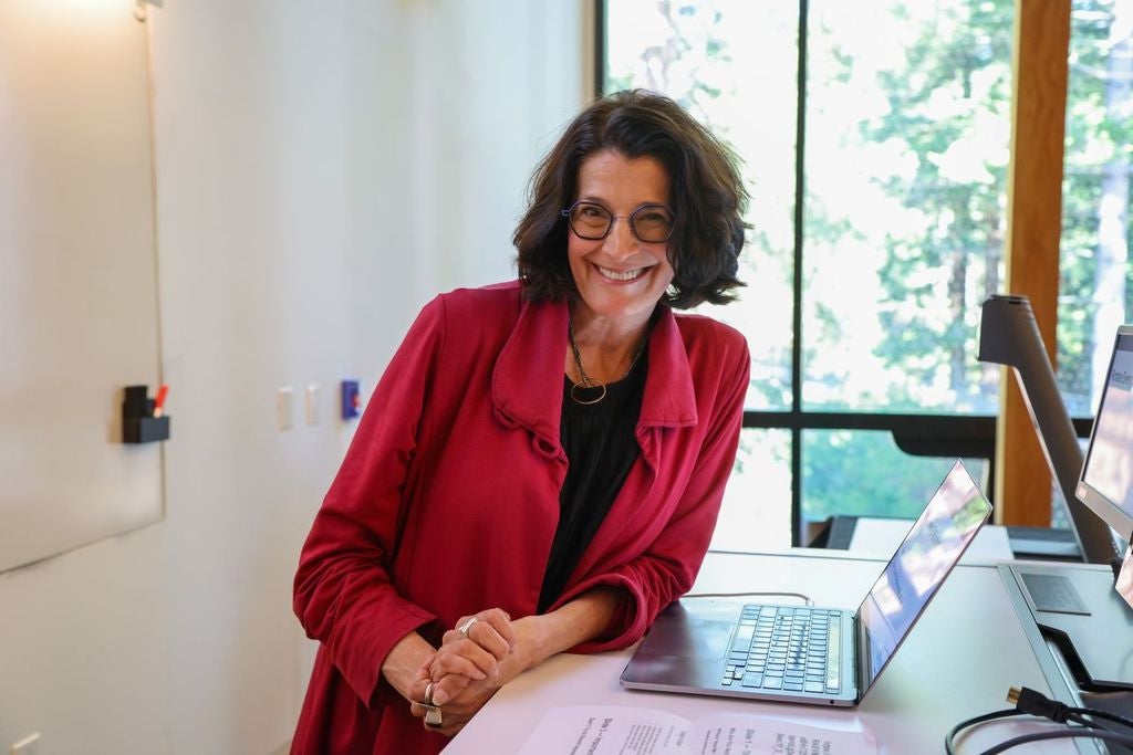 A woman in a red outer shirt and black shirt smiles leaning next to a computer